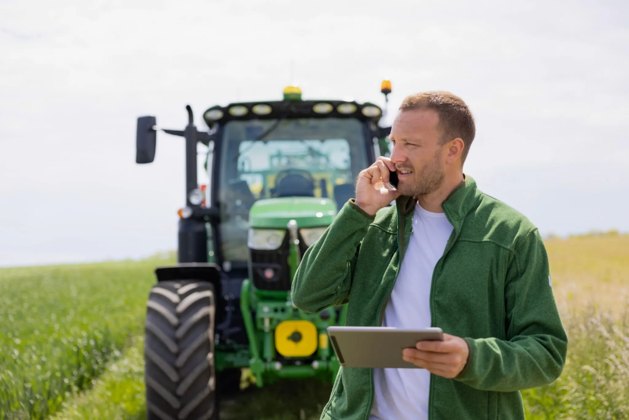 Mann steht am Feld und telefoniert. Im Hintergrund sieht man einen Traktor.